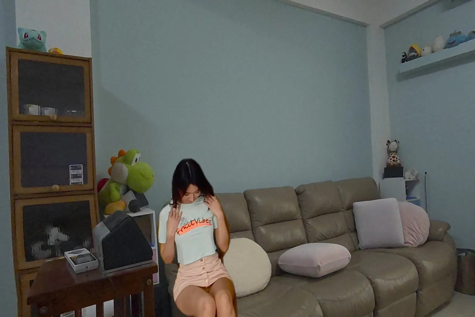 Asiatische Frau in einem hellgrünen T-Shirt und rosa Minirock sitzt auf einem Sofa und zieht leicht an ihrem Shirt, während sie nach unten schaut. Im Hintergrund sind Plüschtiere und Kissen zu sehen.